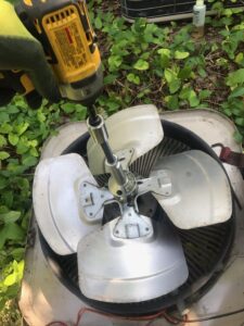 A person with an AC repair background uses a power drill to unscrew the central bolt of a fan on the lawn.